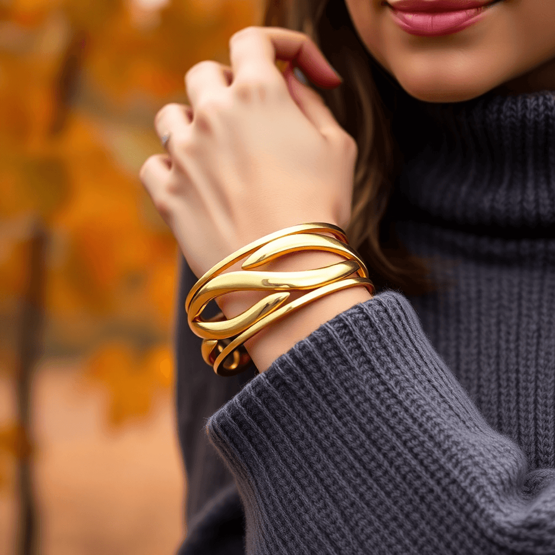 Close-up of a woman wearing a chunky gold cuff bracelet over a cozy cashmere sweater, with blurred autumn leaves in the background.